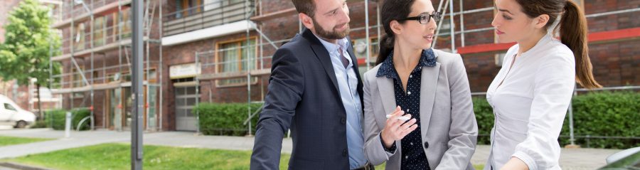 Three business people looking at a blueprint placed on a car bonnet, in front of a building which has a reddish brick façade and is covered in scaffolding, Fast Forward Commercial Excellence