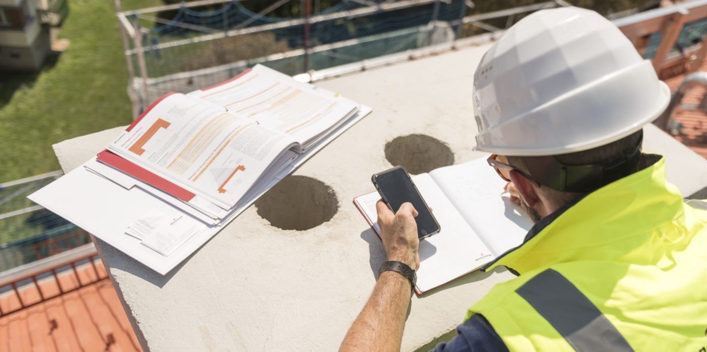 Urban roofer taking notes holding iPhone and writing in an urban location wearing hard hat and safety jacket, Fast Forward Commercial Excellence