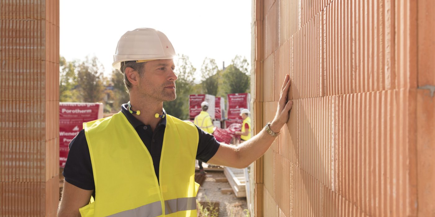 Builder on construction site touching clay block wall, construction workers and clay block pallets in the background