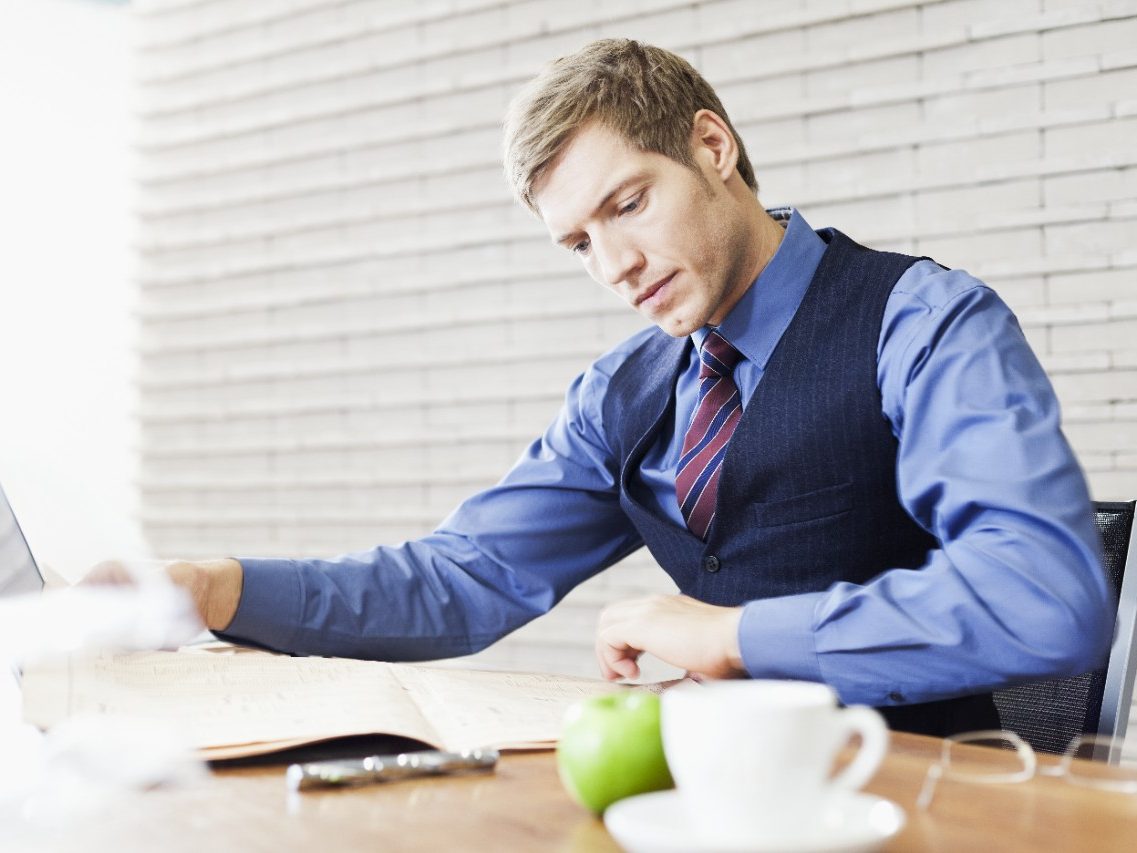 Business man sitting at office desk with laptop comparing financial data from newspaper