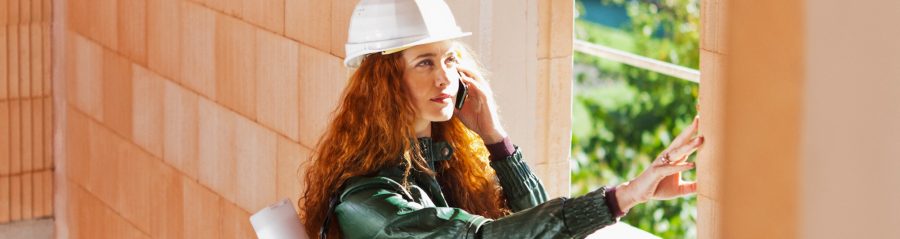 Woman with hard hat and blueprint-casing inside a building shell touching a brick