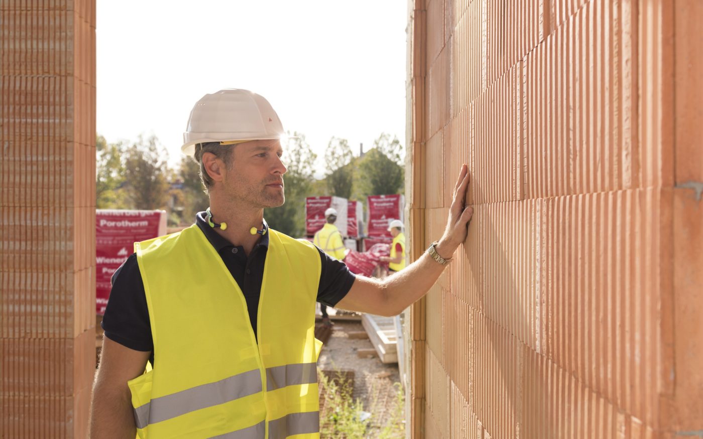 Builder on construction site touching clay block wall, construction workers and clay block pallets in the background