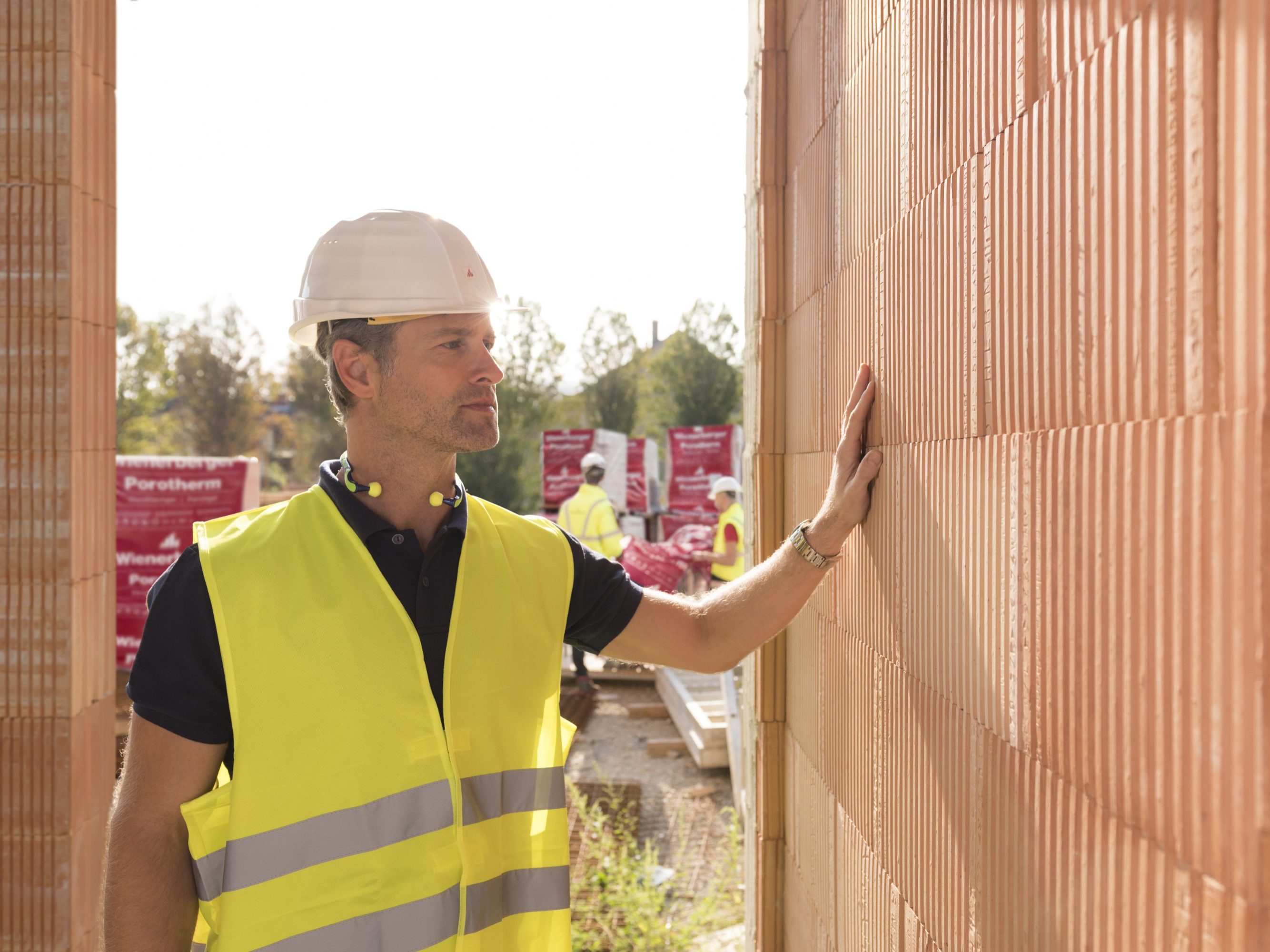 Builder on construction site touching clay block wall, construction workers and clay block pallets in the background