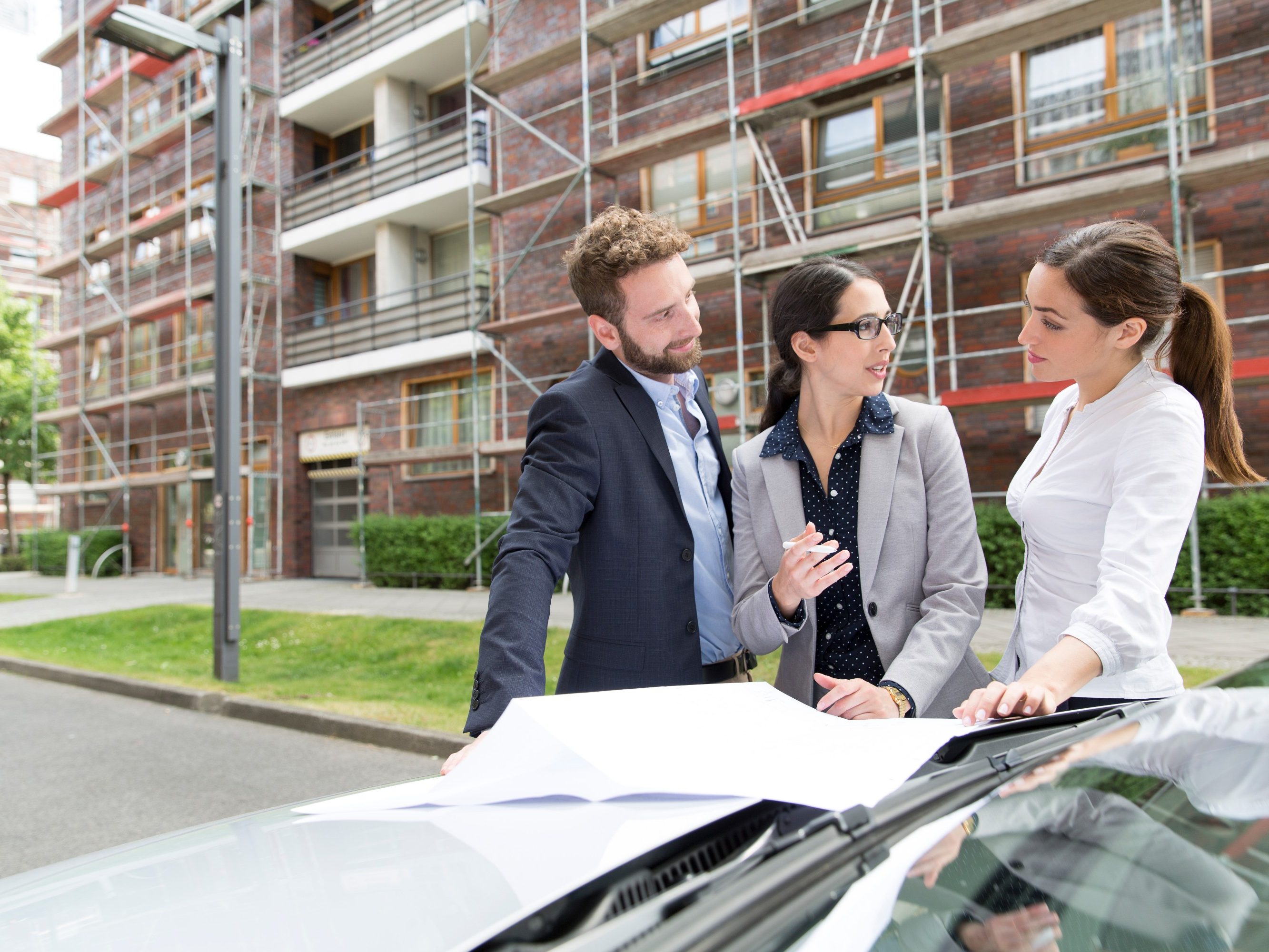 Three business people looking at a blueprint placed on a car bonnet, in front of a building which has a reddish brick façade and is covered in scaffolding, Fast Forward Commercial Excellence