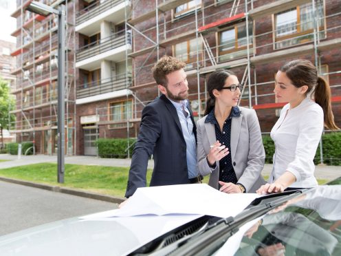 Three business people looking at a blueprint placed on a car bonnet, in front of a building which has a reddish brick façade and is covered in scaffolding, Fast Forward Commercial Excellence