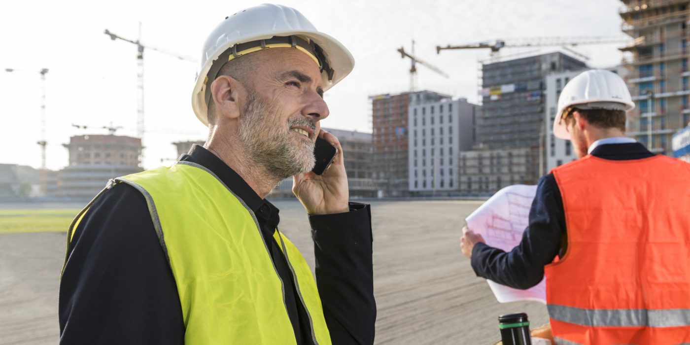 Architect on the phone while his colleague is looking at a plueprint, unfinished buildings in the background, Fast Forward Commercial Excellence