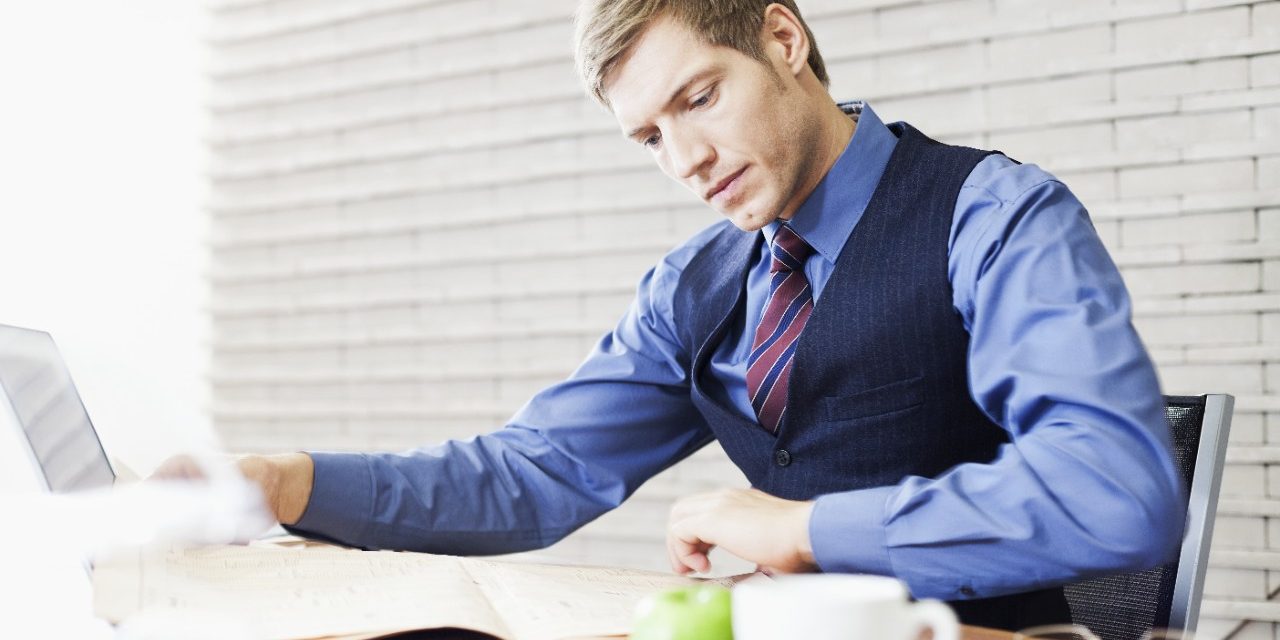 Business man sitting at office desk with laptop comparing financial data from newspaper